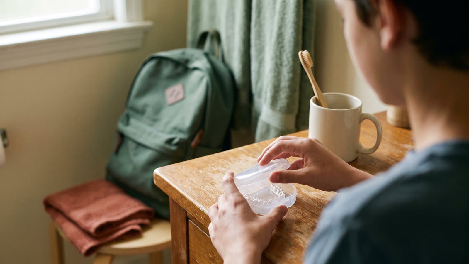 A teen hand closes a retainer case on a bathroom counter with a backpack blurred in the background
